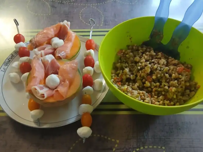 Demi-melons garnis de jambon cru et de billes de mozzarella, brochettes de tomates cerises et mozzarella, à côté d'un saladier de salade