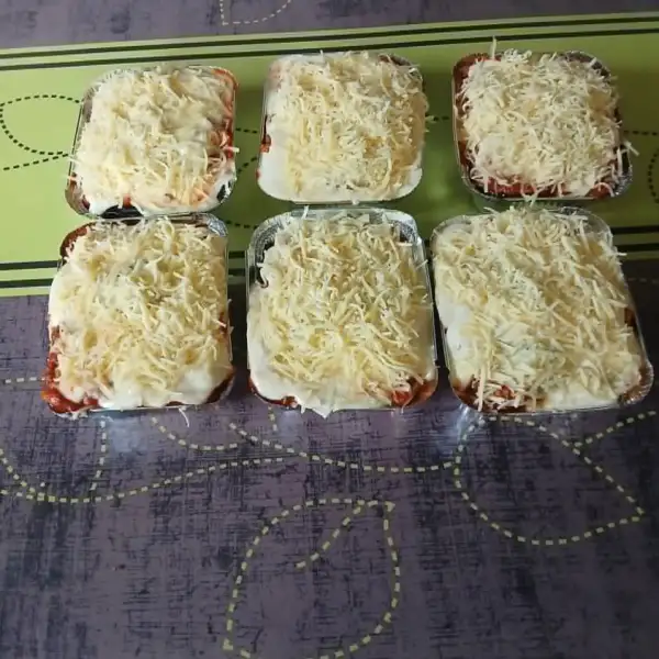 Seis lasañas individuales caseras en charolas, cubiertas de queso rallado antes de hornear